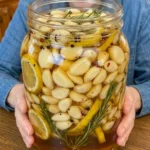 Large jar of honey lemon pickled garlic with rosemary, lemon, peppercorns, and mustard seeds